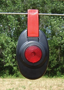 Patriot electric fence light with Red and black traffic light-like device hanging on a wire with green trees in the background